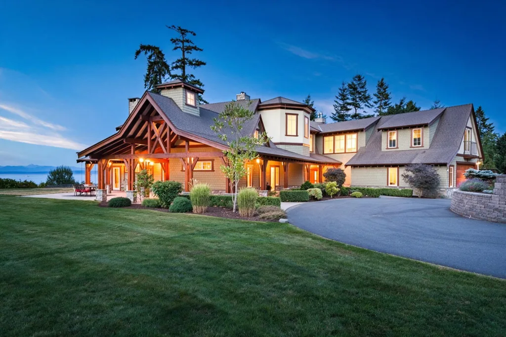 Large, modern house with a lit porch and circular driveway, set on a well-kept lawn at dusk with a lake in the background.