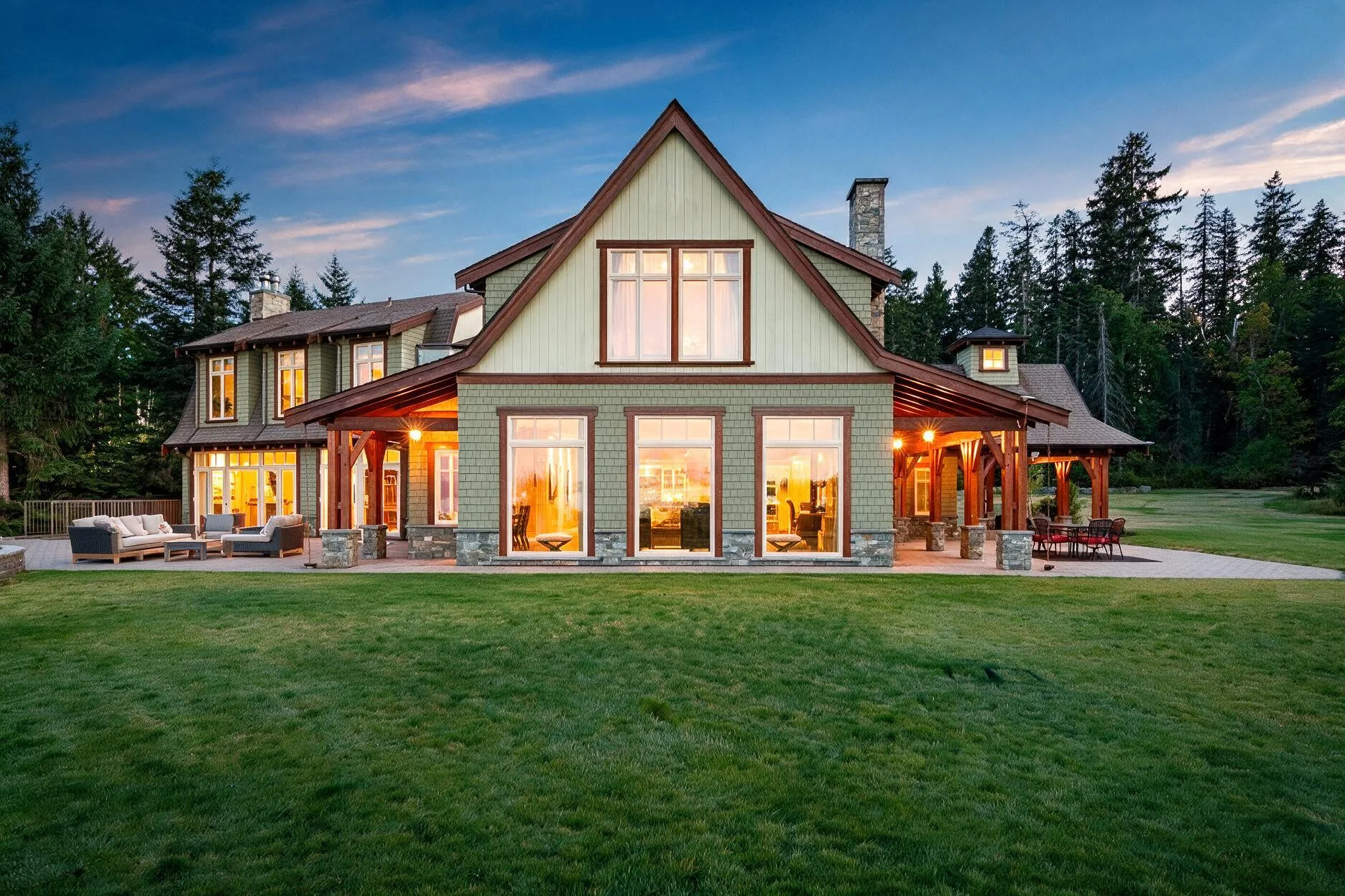 Two-story house with green siding and a prominent peaked roof, large illuminated windows, and a covered patio with outdoor seating at dusk.