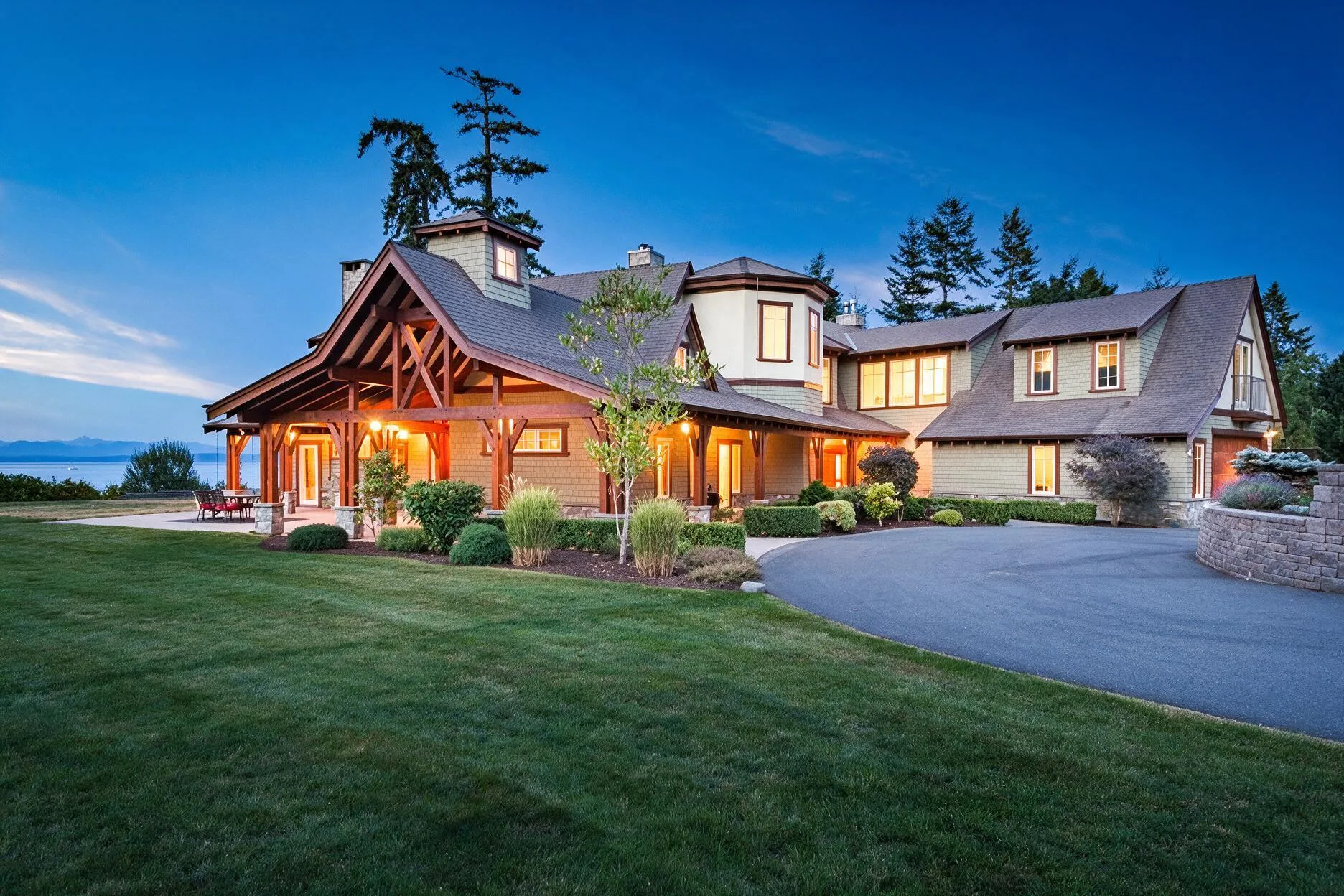 Large, modern house with a lit porch and circular driveway, set on a well-kept lawn at dusk with a lake in the background.